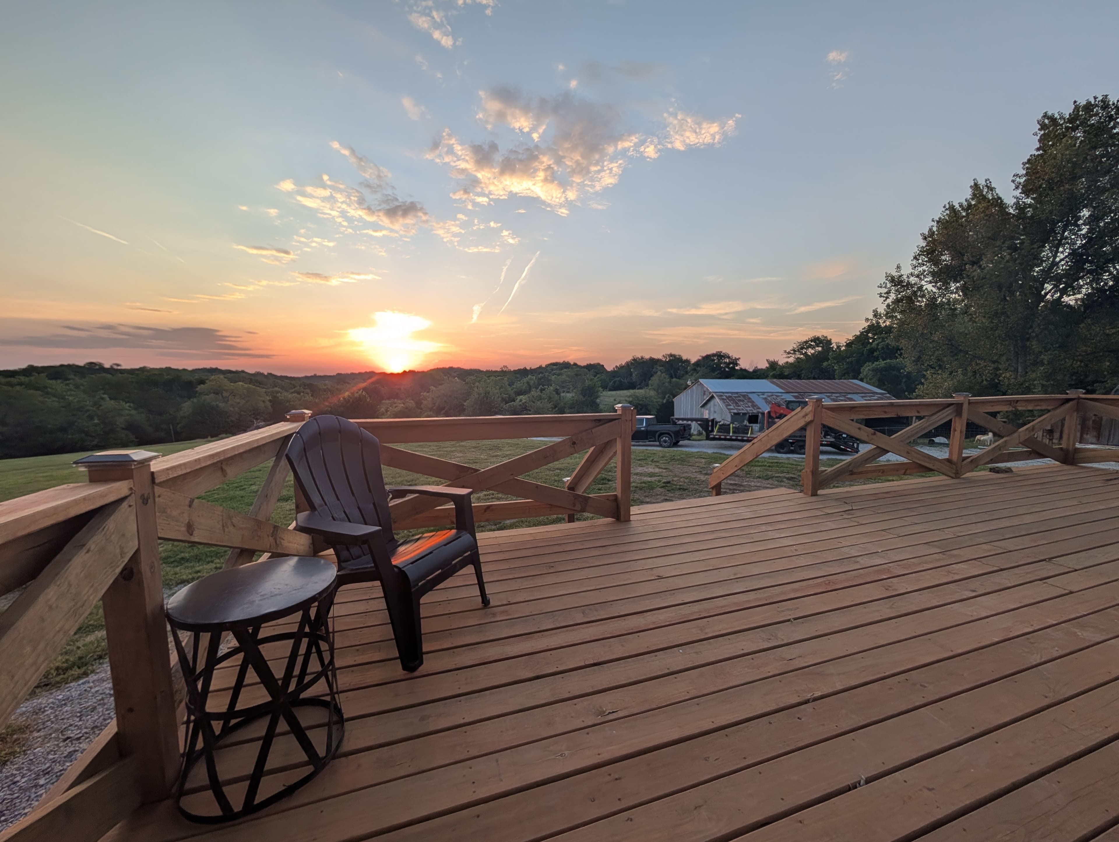 Sunset from the trailer's deck — an Adirondack chair and side table on fresh cedar boards, the barn and timber beyond the railing