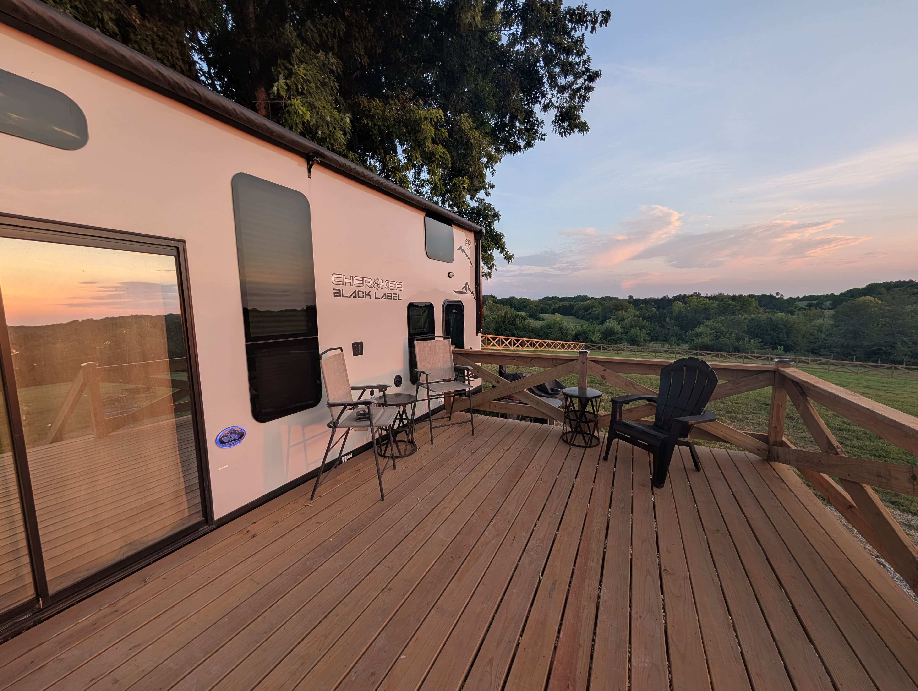 The Cherokee Black Label trailer alongside its wooden deck, chairs set out and the sun setting over the timber and fields beyond