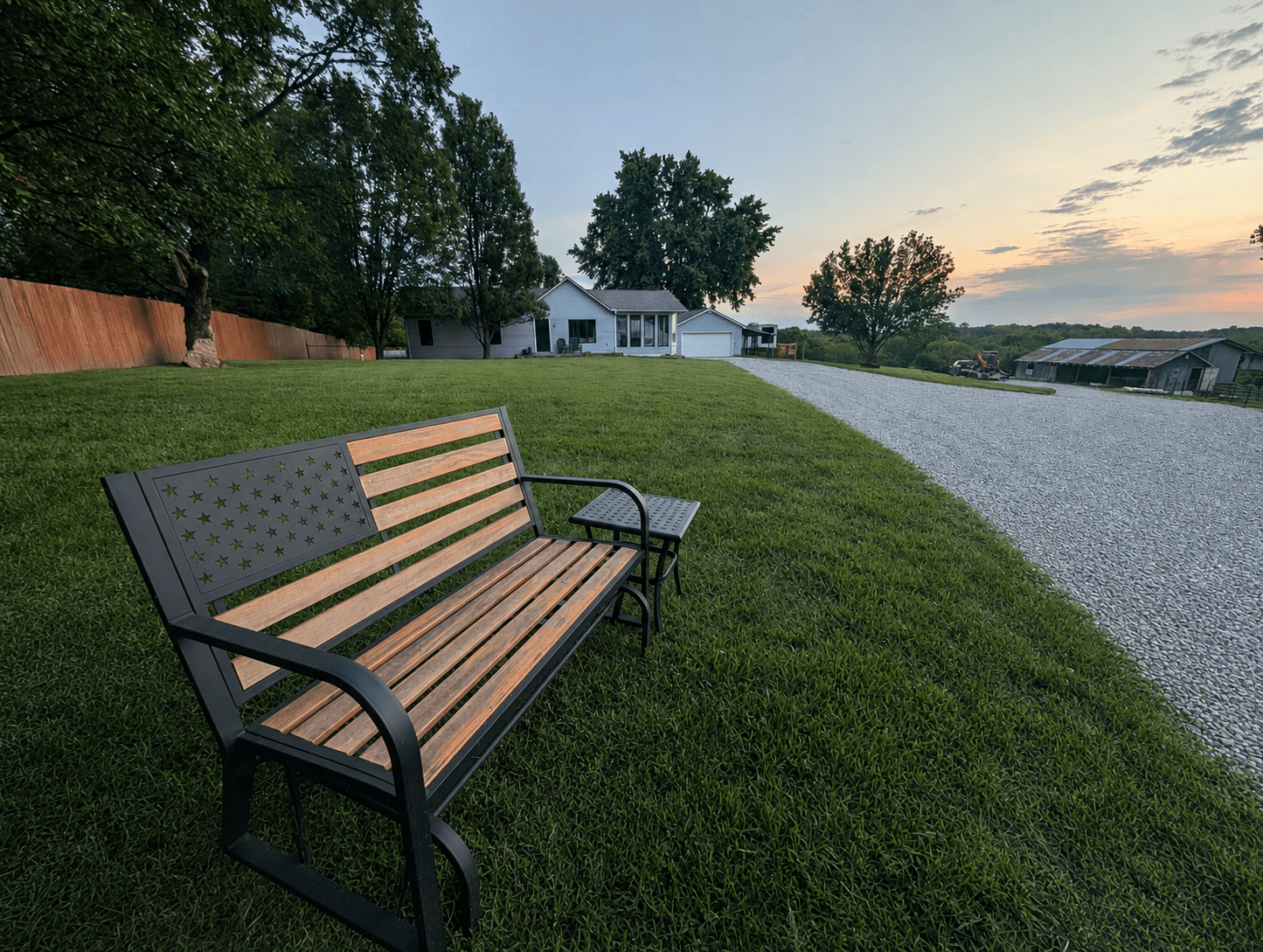 The clubhouse farmhouse across the lawn at sunset, seen from a flag bench beside the gravel drive, with the barn off to the right