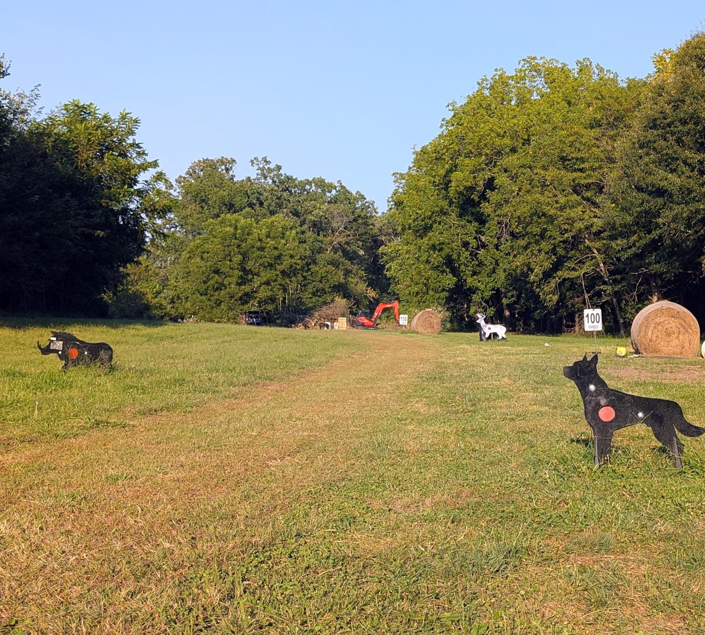 Looking downrange along the mowed rifle line — steel animal silhouettes and a white deer target staged at marked yardages, with round hay bale backstops against the treeline